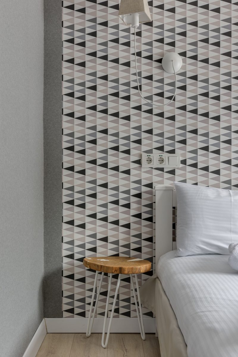 Single geometric triangle-pattern wallpaper feature wall in an otherwise neutral bedroom with a wooden stool and wall lamp