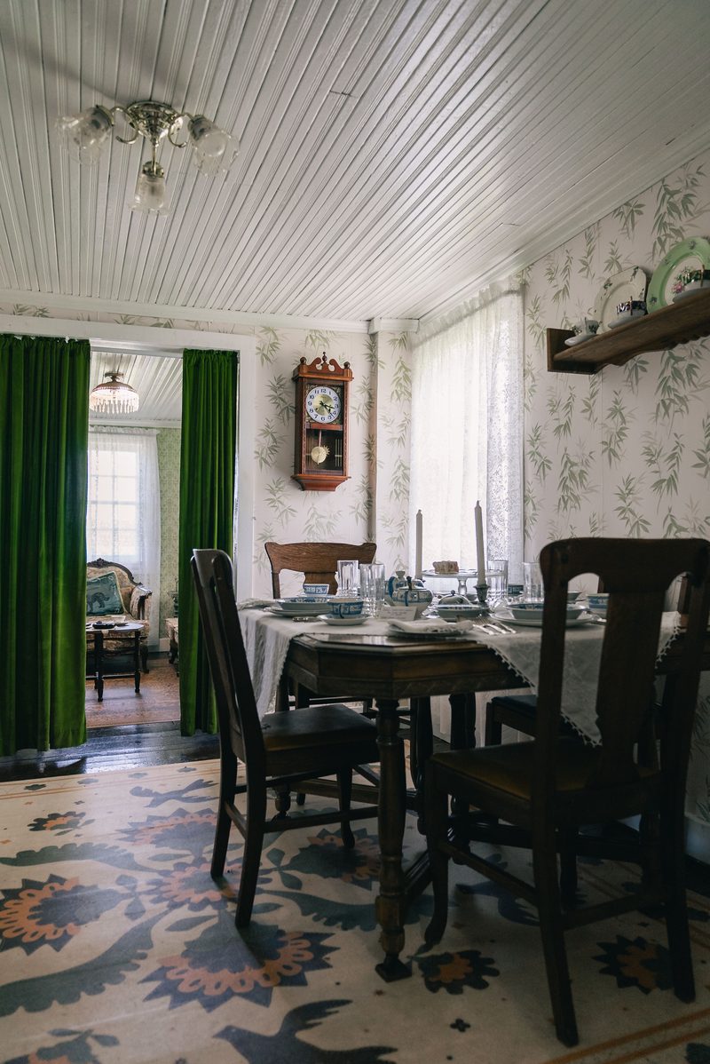 Traditional dining room with a set table, antique furniture, and patterned wallpaper framed by green drapery