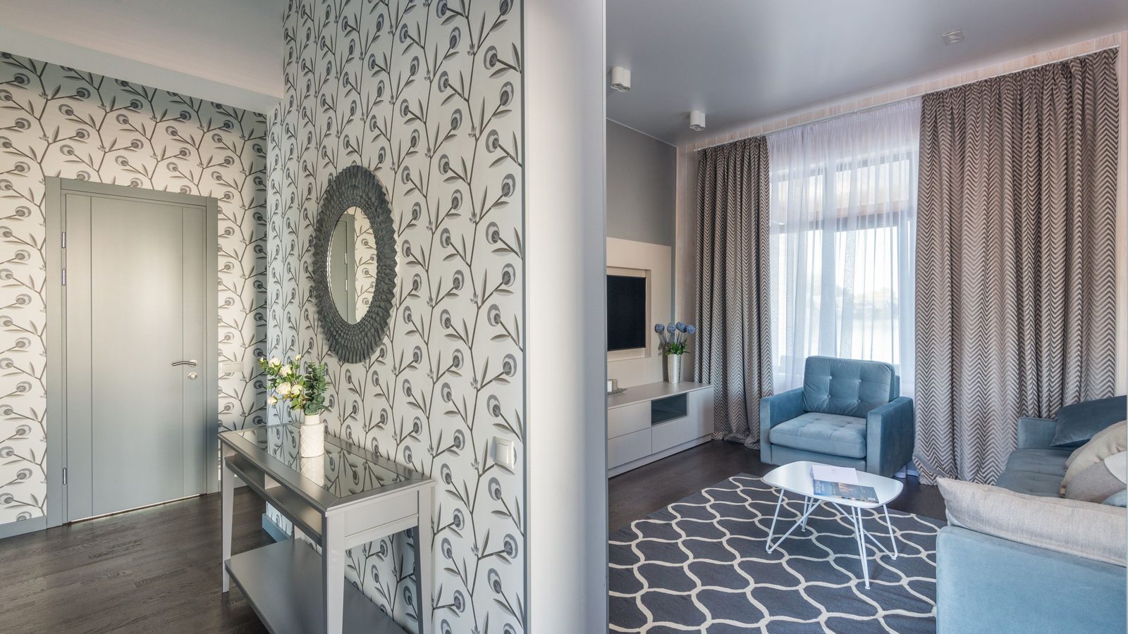 Sunlit modern living room with a soft grey-green botanical wallpaper feature wall, round mirror, console table and blue armchairs