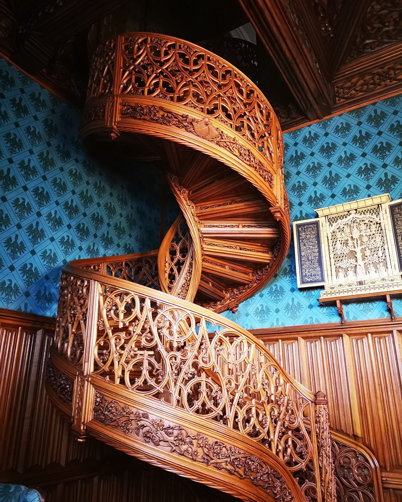 Carved wooden spiral staircase in a detailed interior with patterned wallpaper — a study in careful, precise craftsmanship